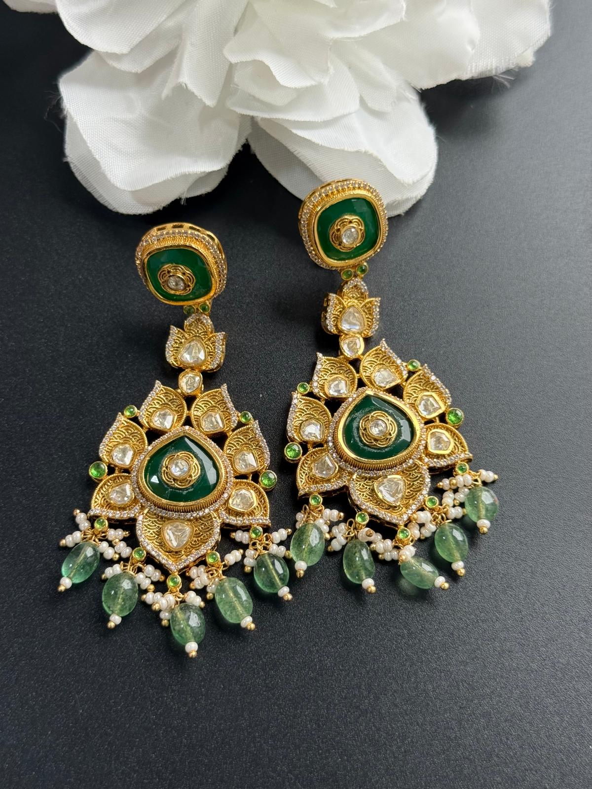 Ornate gold and green earrings on a dark surface with white flowers in the background