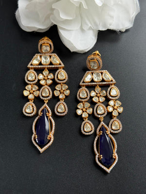 Pair of gold and blue earrings on a dark surface with white flowers in the background