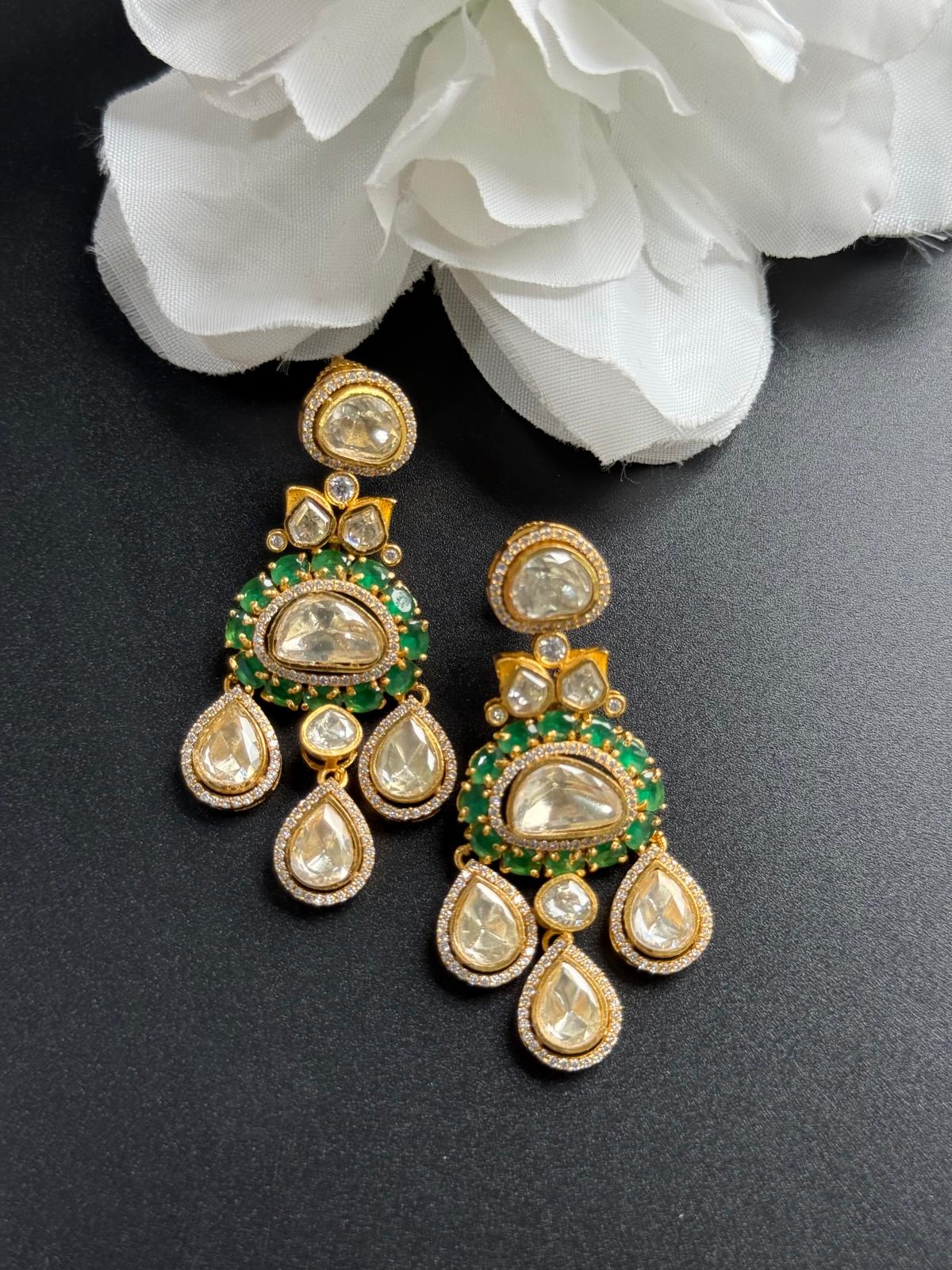 Ornate earrings with green and gold accents on a dark surface with white flowers.