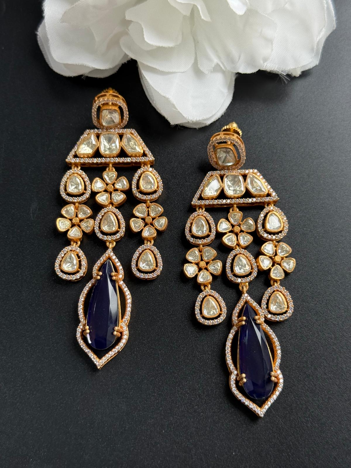 Pair of gold and blue earrings on a dark surface with white flowers in the background